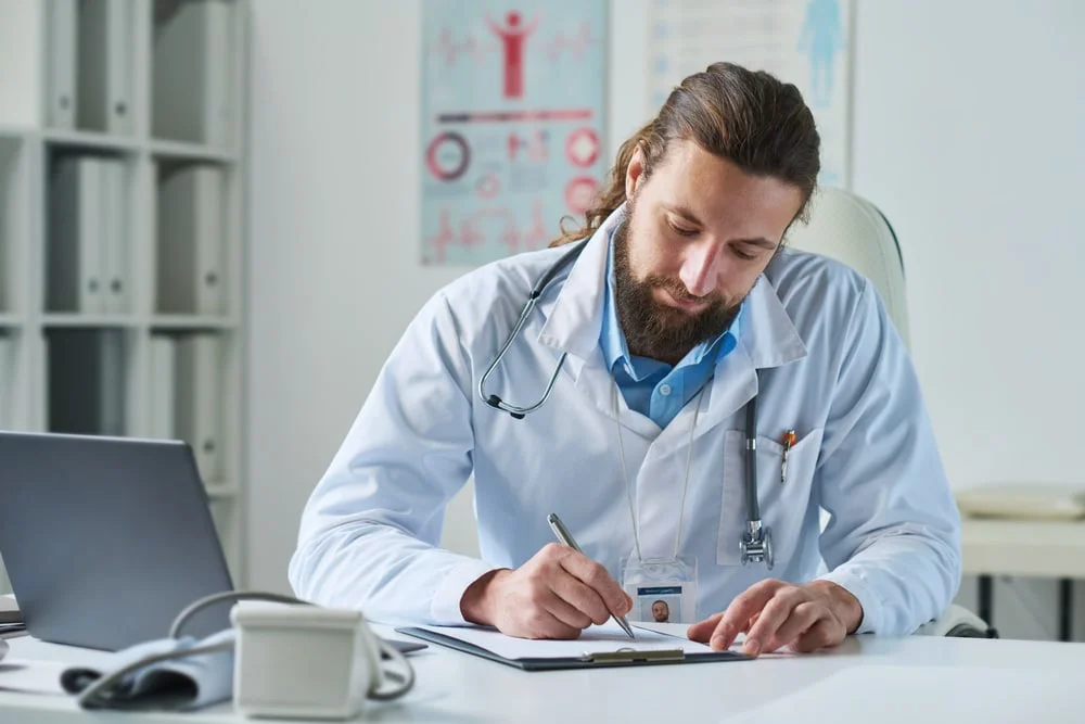 Young bearded male clinician in lab coat making notes in medical document while sitting by workplace in hospital